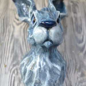 May include: A blue ceramic sculpture of a rabbit's head with a textured surface. The rabbit has a serious expression and blue eyes.