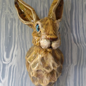 May include: A ceramic hare wall sculpture with a detailed, textured surface. The hare has large, upright ears, blue eyes, and a light brown and tan colour scheme. The sculpture is mounted on a light blue and white wooden background.