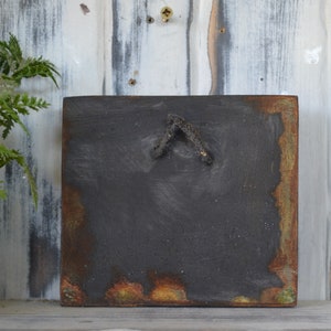 May include: A square, black, wooden sign with a rusty, green, and orange border. The sign has a small, black, metal hook on the back.