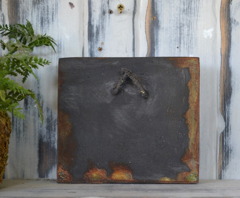 May include: A square, black, wooden sign with a rusty, green, and orange border. The sign has a small, black, metal hook on the back.