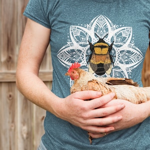 May include: A teal t-shirt with a bumblebee graphic inside a white mandala design. A person holds a brown and white chicken. The background is a wooden fence.