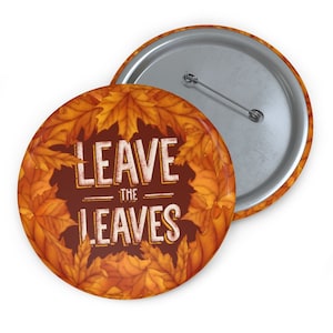 May include: A round button with a fall theme. The button features the text "LEAVE THE LEAVES" in white, surrounded by a border of orange and yellow autumn leaves. The back of the button is silver with a pin.