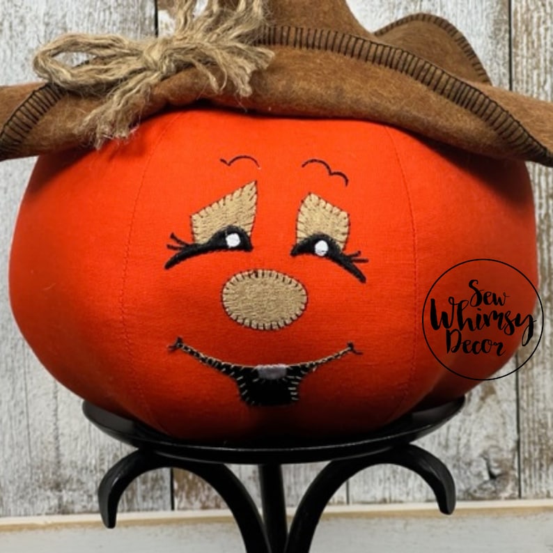 Pu&ograve; includere: Una zucca di stoffa arancione con un volto sorridente, un cappello marrone e un fiocco di rafia. La zucca &egrave; appoggiata su un supporto di metallo nero. La zucca ha un volto cucito con ciglia nere e una bocca cucita. Il testo "Sew Whimsy Decor" &egrave; sul lato destro.