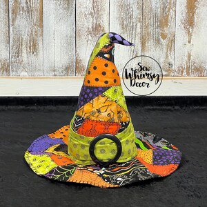 May include: A fabric witch hat featuring a patchwork design of orange, lime green, and purple patterns including skeletons, spiderwebs, and polka dots. The hat has a wide brim and a lime green band with a large black circular buckle at the center.