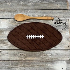 May include: A brown football-shaped trivet with white stitching detail. A wooden spoon rests above the trivet. The image is on a rustic, weathered wood surface. The text "Sew Whimsy Decor" is in a circle.