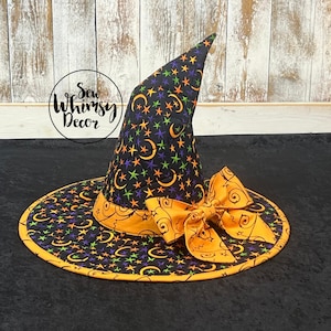 May include: A quilted witch hat featuring a black fabric base with a pattern of orange and purple crescent moons and green stars. The hat includes an orange band and a large orange bow with black swirling line designs. Text reads Sew Whimsy Decor.