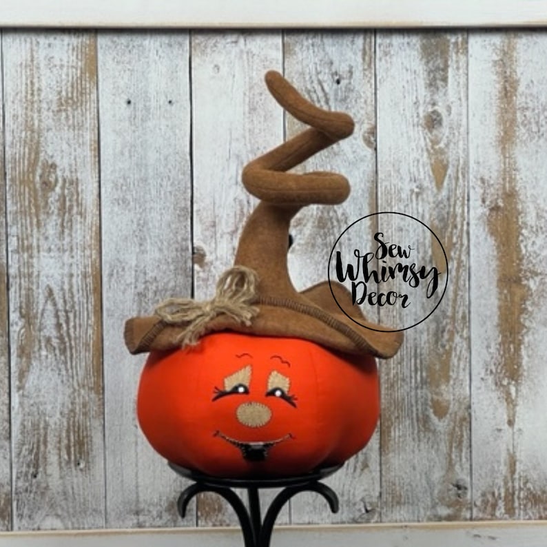 Pu&ograve; includere: Una zucca di stoffa arancione con un volto sorridente, che indossa un cappello da strega marrone con la parte superiore riccia. La zucca &egrave; su un supporto di metallo nero. Il testo "Sew Whimsy Decor" &egrave; in un cerchio a destra.