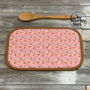 May include: A pink quilted hot pad with a brown border, featuring a pattern of gingerbread men and candy canes. A wooden spoon rests above the pad. The image includes the text "Sew Whimsy Decor".