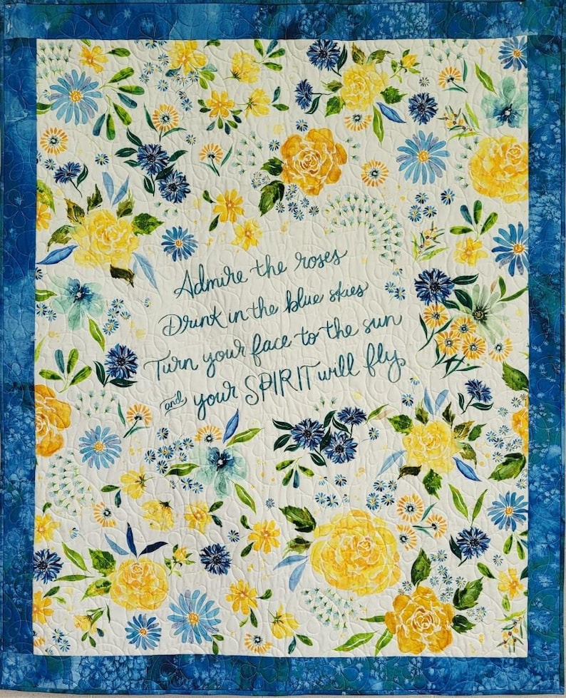 May include: A floral quilt with a white background, featuring yellow and blue flowers and green leaves. The quilt has a blue border and includes the text "Admire the roses, Drink in the blue skies, Turn your face to the sun and your SPIRIT will fly."