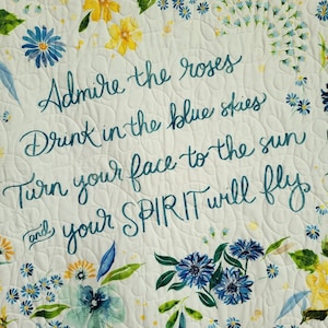 May include: A white floral quilt with yellow and blue flowers and green leaves. The quilt features the text "Admire the roses, Drink in the blue skies, Turn your face to the sun and your SPIRIT will fly" in teal script.