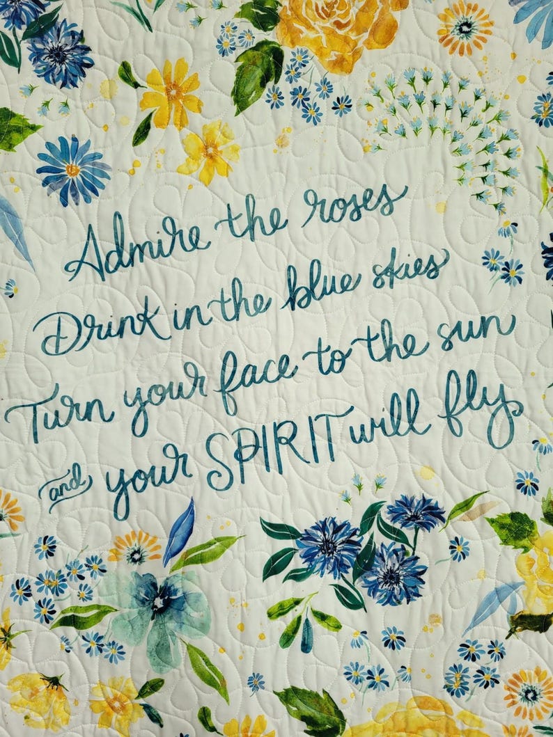 May include: A white floral quilt with yellow and blue flowers and green leaves. The quilt features the text "Admire the roses, Drink in the blue skies, Turn your face to the sun and your SPIRIT will fly" in teal script.