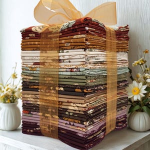 May include: A tall stack of folded fabric squares tied together with a translucent gold ribbon. The fabric pieces feature various autumn-inspired patterns in shades of deep red, burnt orange, mustard yellow, cream, sage green, brown, and plum.