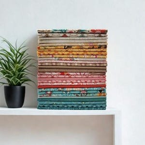 May include: A stack of colorful fabric squares with various floral and geometric patterns. The fabrics are in shades of pink, yellow, blue, and brown. A small potted plant sits to the left of the fabric stack on a white shelf.