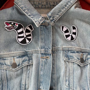 Puede incluir: Una chaqueta vaquera azul claro con dos parches en blanco y negro. Los parches son de una criatura rayada con boca y dientes.