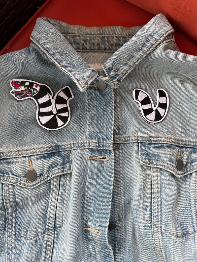 Puede incluir: Una chaqueta vaquera azul claro con dos parches en blanco y negro. Los parches son de una criatura rayada con boca y dientes.