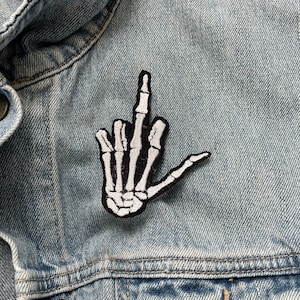 May include: A white skeleton hand giving the middle finger, embroidered on a blue denim jacket.