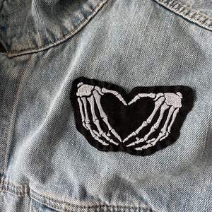 May include: A denim jacket with a black and white embroidered patch of two skeleton hands forming a heart shape.