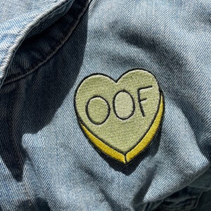 May include: A light green heart-shaped patch with yellow stitching and the word "OOF" embroidered in white.