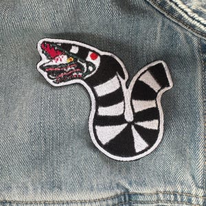Puede incluir: Un parche de rayas blancas y negras de una criatura con boca roja y ojos amarillos. El parche est&aacute; cosido en una chaqueta vaquera azul.
