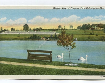 May include: A vintage postcard showing a general view of Firestone Park in Columbiana, Ohio. The scene features a blue lake with two white swans, a wooden bench on the grassy bank, a small tree, and a long white building in the distance.