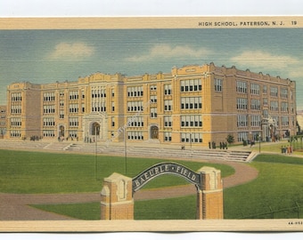 May include: A vintage postcard showing a large, multi-story yellow brick high school building in Paterson, N.J. In the foreground, a curved metal archway with the text BAEURLE FIELD stands over a green lawn and a dirt track.