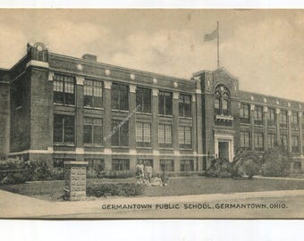May include: A vintage postcard featuring a multi-story brick building identified as Germantown Public School, Germantown, Ohio. The structure has numerous rectangular windows, a central entrance, and an American flag flying from the roof.