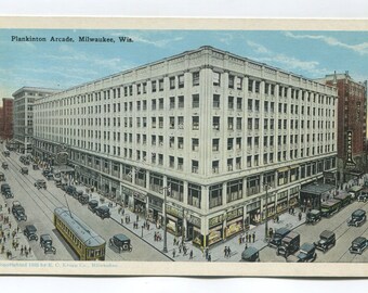 May include: A vintage postcard showing the Plankinton Arcade in Milwaukee, Wisconsin. The large, multi-story building occupies a street corner, surrounded by early 20th-century automobiles, a yellow streetcar, and pedestrians on the sidewalk.