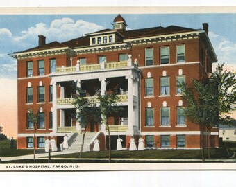 May include: A vintage postcard featuring a multi-story red brick building with a white columned porch and balcony. The text at the bottom reads ST. LUKE'S HOSPITAL, FARGO, N. D. Several figures in white uniforms stand near the front entrance.