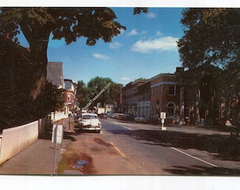 May include: A vintage postcard view of a town street featuring a white picket fence on the left, a white car parked along the curb, and multi-story brick buildings lining the road under a clear blue sky with scattered clouds and green trees.