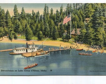 May include: A vintage postcard showing Sweyolakan on Lake Coeur d'Alene, Idaho. A white steamboat is docked at a wooden pier near a sandy beach. A large American flag flies near a forest of pine trees. Several small rowboats are on the water.