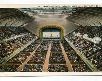 May include: Vintage postcard of the interior of the Cleveland Public Hall in Cleveland, Ohio. The image shows a large auditorium with rows of seating filled with people. A stage is visible at the far end, with a decorative arch and a performance area.