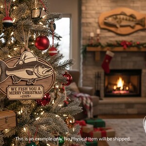 May include: Wooden Christmas ornament featuring a fish wearing a Santa hat. The ornament reads "We Fish You a Merry Christmas - 2025." The ornament is hanging on a decorated Christmas tree with other ornaments and lights.