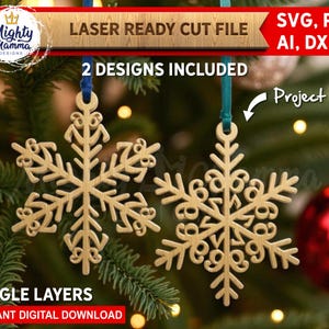 May include: Two laser-cut wooden snowflake decorations suspended by blue and teal ribbons, set against a backdrop of Christmas tree branches and red baubles. The image includes text that reads "LASER READY CUT FILE" and "2 DESIGNS INCLUDED".