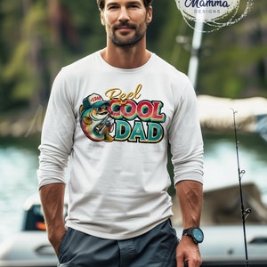 Peut inclure: Un t-shirt blanc &agrave; manches longues avec un motif color&eacute; d'un poisson portant des lunettes de soleil et un chapeau qui dit "#1 DAD". Le motif comprend &eacute;galement le texte "Reel Cool Dad".