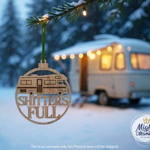 May include: A wooden Christmas ornament with a camper design and the words "SHITTERS FULL". The ornament is hanging from a green ribbon against a snowy winter backdrop. A vintage camper is in the background.
