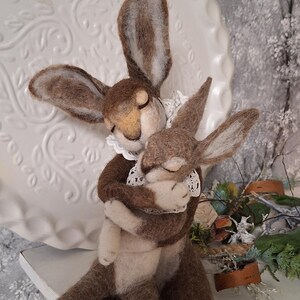 Könnte beinhalten: Zwei Filzhasen in Braun und Weiß umarmen sich. Der größere Hase hat lange Ohren und einen weißen Spitzenkragen. Der kleinere Hase ist in seinen Armen eingebettet. Der Hintergrund umfasst einen weißen Zierteller und eine winterliche Szene.