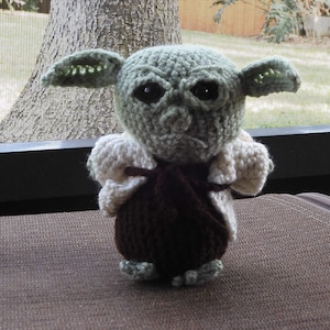 May include: A crocheted green and brown Yoda doll wearing a white crocheted jacket. The doll has large black eyes and a serious expression.