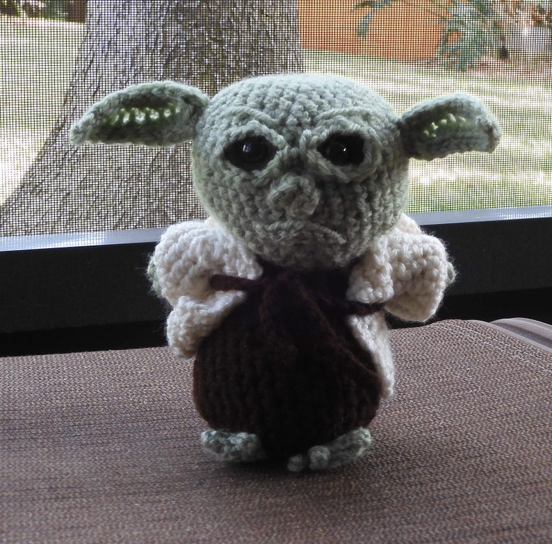 May include: A crocheted green and brown Yoda doll wearing a white crocheted jacket. The doll has large black eyes and a serious expression.
