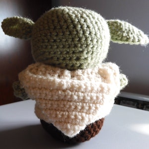 May include: A crocheted green and brown plush toy with large ears. The toy is wearing a white crocheted robe.
