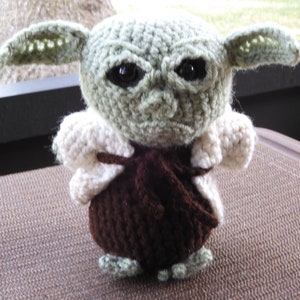 May include: A crocheted plush toy of a green alien character with large ears, brown clothing, and white fur trim. The toy has black eyes and a serious expression.