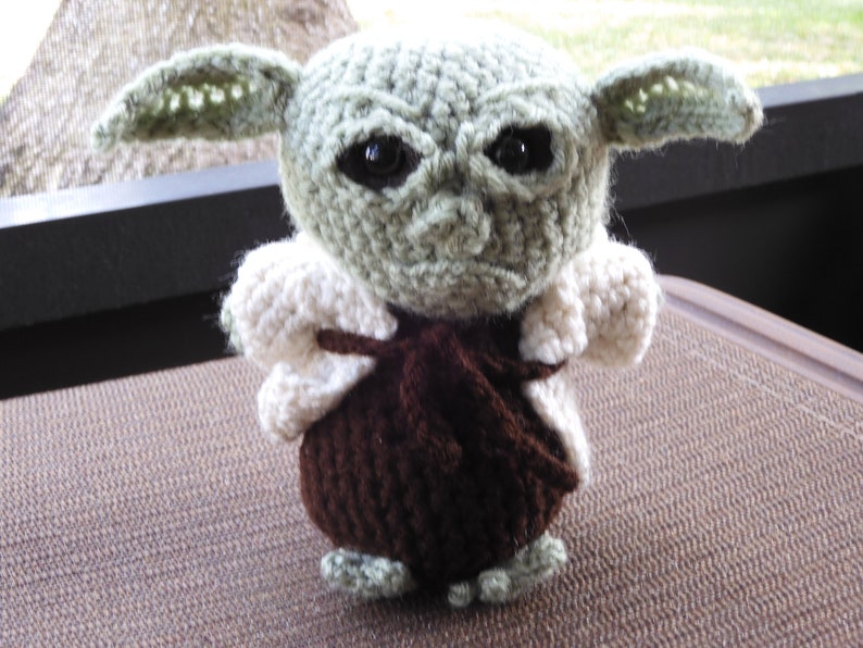 May include: A crocheted plush toy of a green alien character with large ears, brown clothing, and white fur trim. The toy has black eyes and a serious expression.