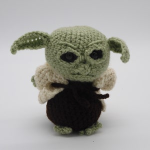 May include: A crocheted plush toy of a green character with large ears, brown clothing, and black eyes.