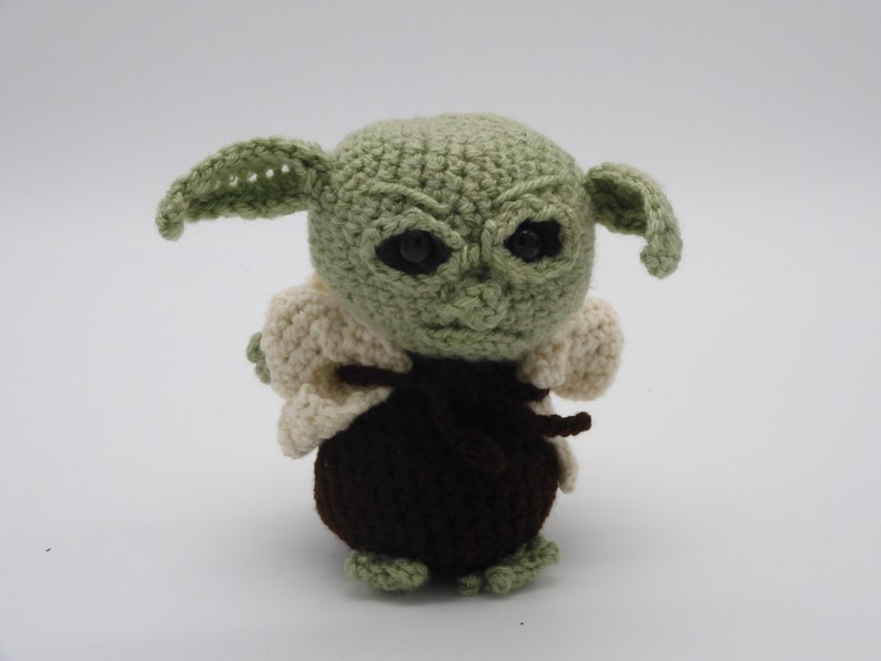 May include: A crocheted plush toy of a green character with large ears, brown clothing, and black eyes.