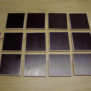 Könnte beinhalten: Zwölf quadratische Untersetzer mit dunkler Oberfläche und hellem Holzrand, angeordnet in drei Reihen. Die Untersetzer sind auf einer Holzoberfläche platziert. Jeder Untersetzer misst etwa 10 cm x 10 cm.