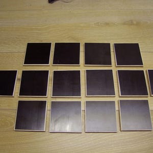 May include: A set of 15 black square coasters. The coasters are arranged in a pyramid shape on a light wood surface.