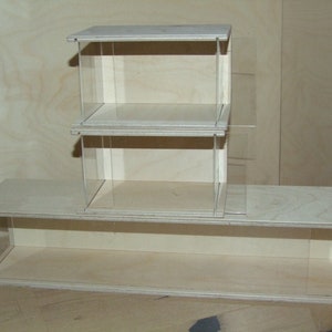 May include: A three-tiered wooden display case with clear acrylic sides and shelves. The case is made of light wood and has a natural finish.