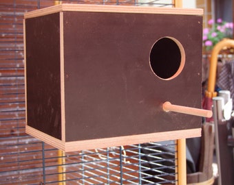 Könnte beinhalten: Ein braunes Vogelhaus aus Holz mit einem runden Eingangsloch und einem kleinen Holz-Sitzplatz. Das Vogelhaus ist an einem Metallkäfig befestigt.