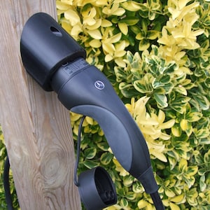 May include: A black and gray electric vehicle charging connector with a Mercedes-Benz logo. The charger is mounted on a wooden post, with a backdrop of green and yellow foliage.