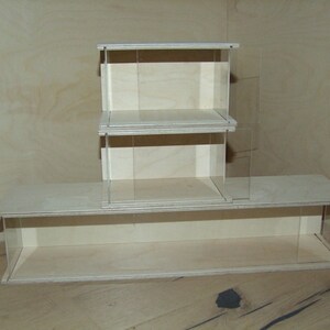 May include: A three-tiered wooden display shelf with clear acrylic sides. The shelf is made of light wood and has a simple, modern design.