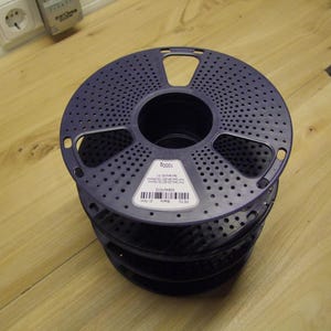 Könnte beinhalten: Ein Stapel dunkelblauer 3D-Drucker-Filamentrollen. Jede Rolle ist rund mit einer Reihe kleiner Löcher und einer zentralen Öffnung. Ein Etikett mit Text und einem Barcode ist auf der obersten Rolle sichtbar. Die Rollen sind auf einer hellen Holzoberfläche gestapelt.